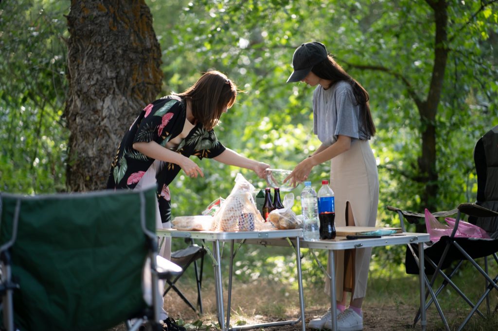 people prepare food for a picnic in wooded area. standing near a table with food and drinks